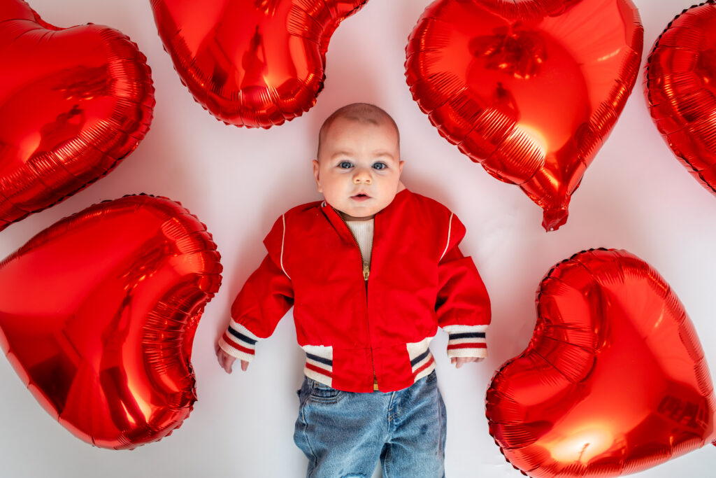 Cute baby in red jacket laying amongst red heart balloons
